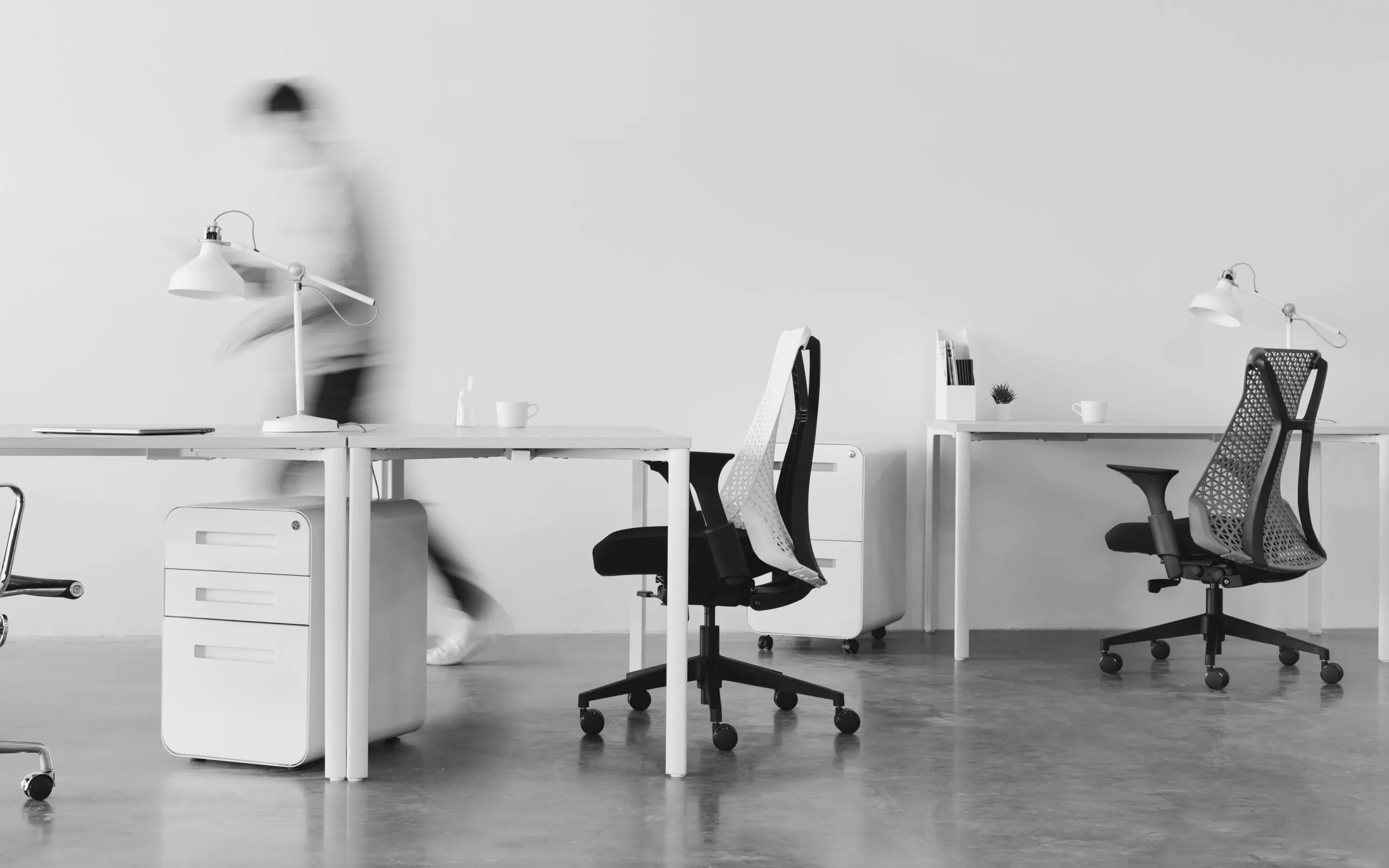 Black and white image of an office with tables and chairs, while a person walks by.